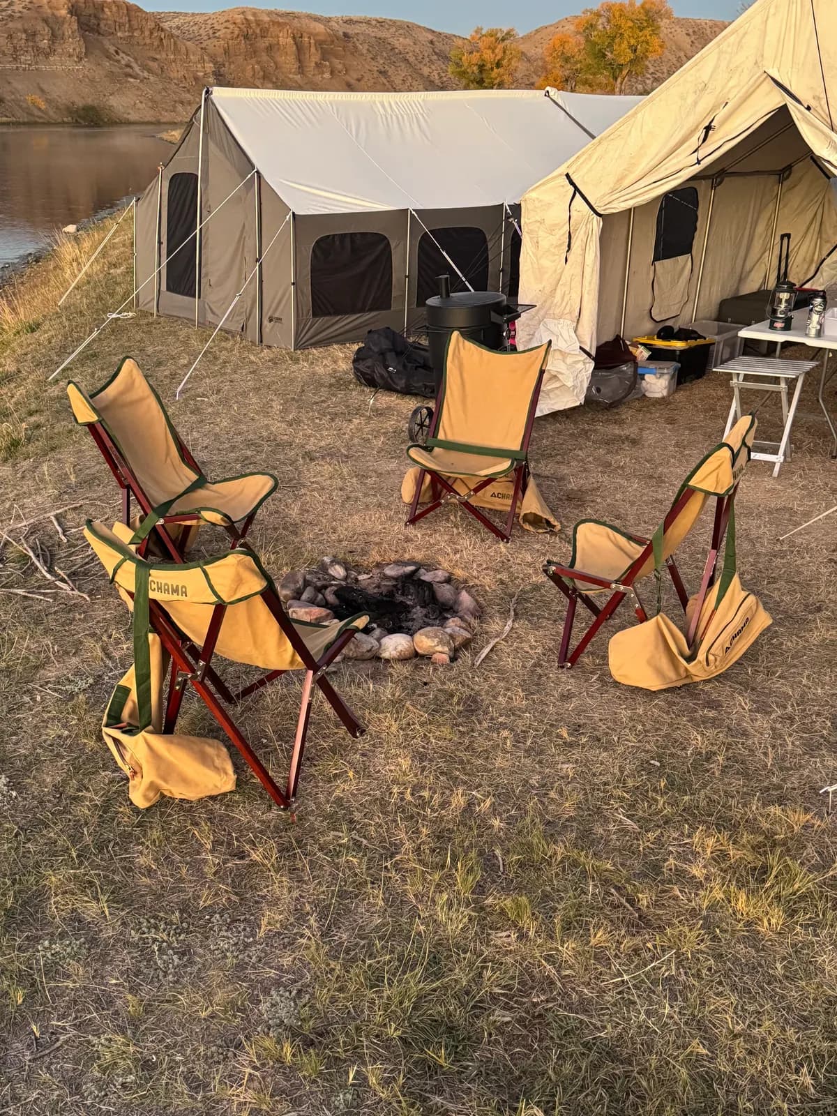 Canvas camp chairs around a stone fire ring in front of two canvas tents on a riverbank.
