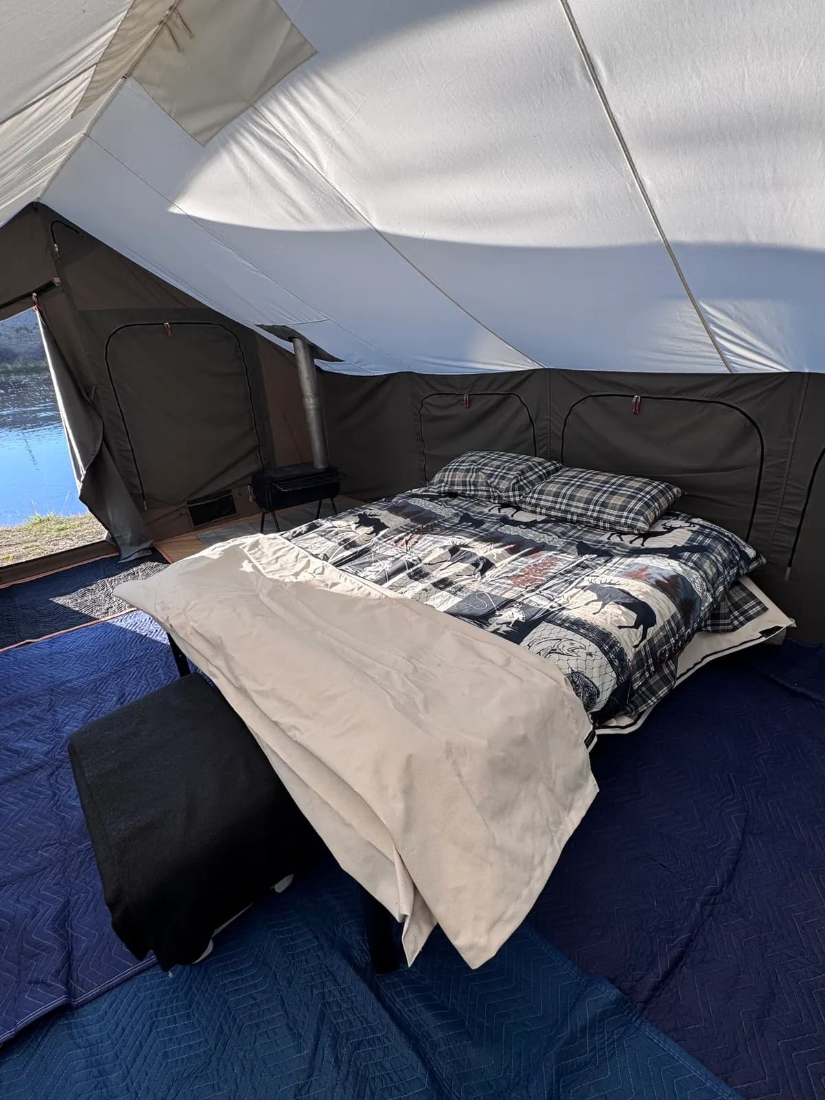 Inside a canvas cabin tent: a made queen bed with plaid pillows, a small wood stove with its pipe through the roof, and an open door looking out on a river.